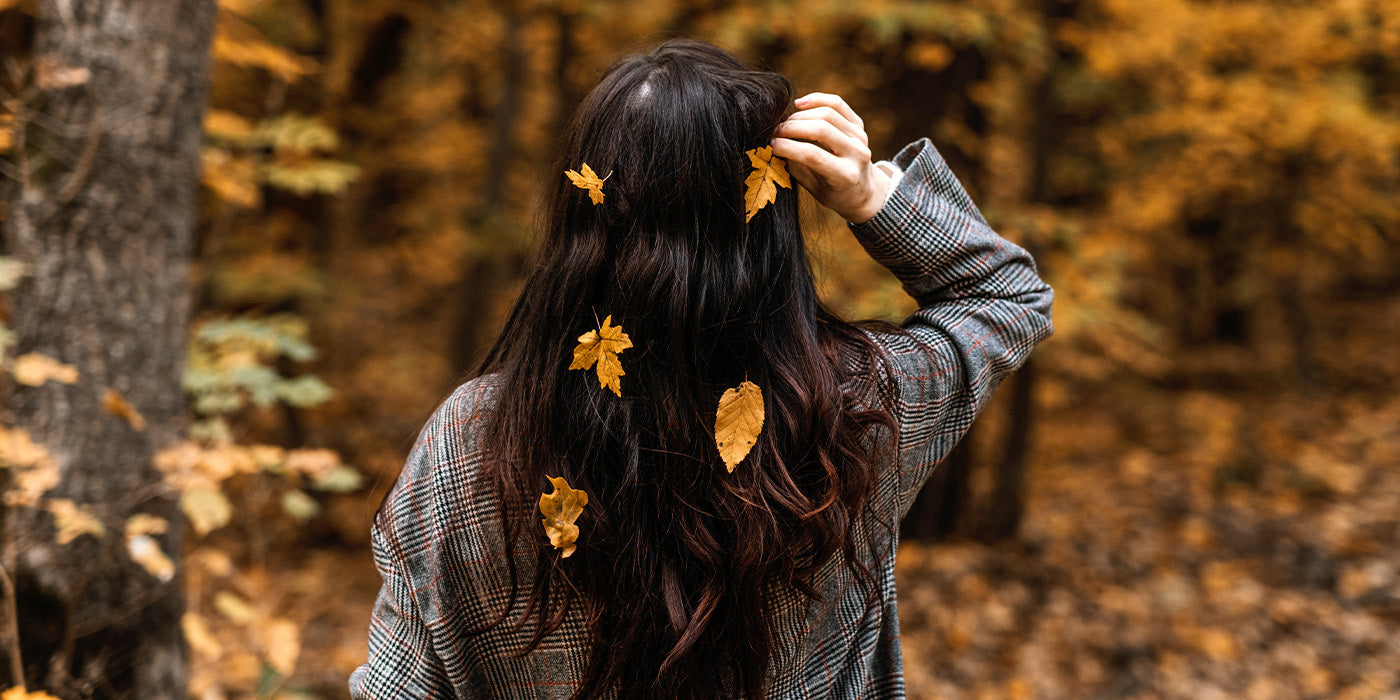 Frau mit langen brauen Haaren steht draußen im Wald und Laub ist in ihren Haaren
